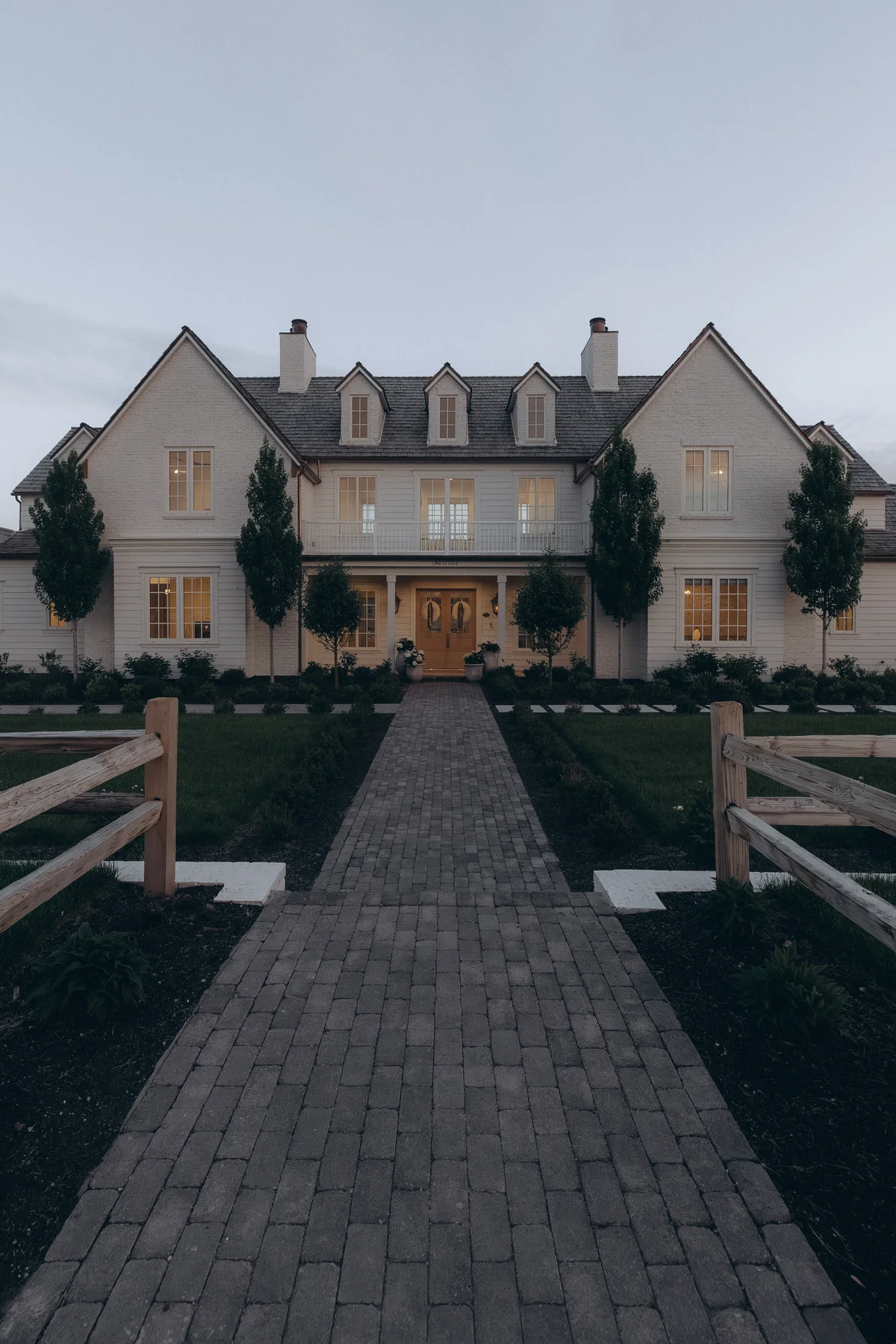 Symmetrical white farmhouse front elevation at dusk with dormers and split-rail fence